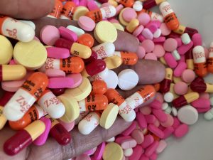 Eine Hand voller Medikamente und unter der Hand ist eine Fläche zu sehen mit ebenfalls verschiedensten Pillen in orange, rosa, gelb, weiß und rot.